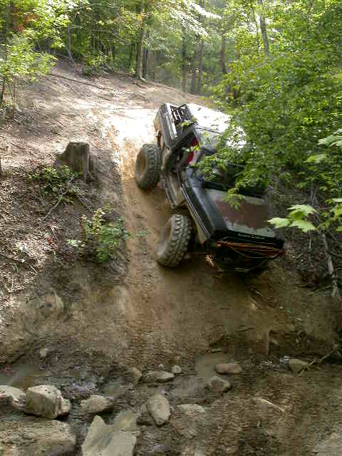 1990 Ford Bronco - A black SUV navigates a challenging off-road trail, climbing a steep, muddy incline surrounded by trees.