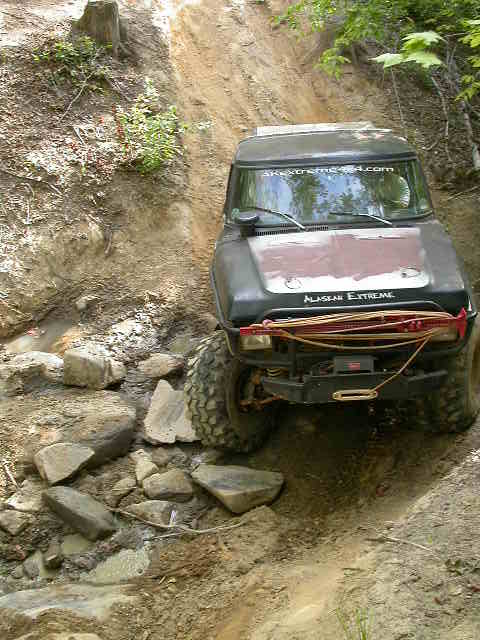 1990 Ford Bronco - This modified off-road vehicle navigates a challenging, rocky incline on a wooded trail.