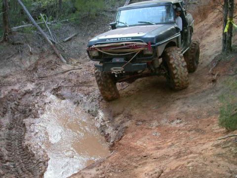 1990 Ford Bronco - Off-road vehicle navigating a muddy trail with a deep water-filled rut.