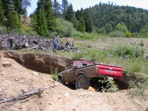 1988 Ford Bronco - A red Toyota pickup truck maneuvers through a challenging off-road terrain, with two people observing in the background.