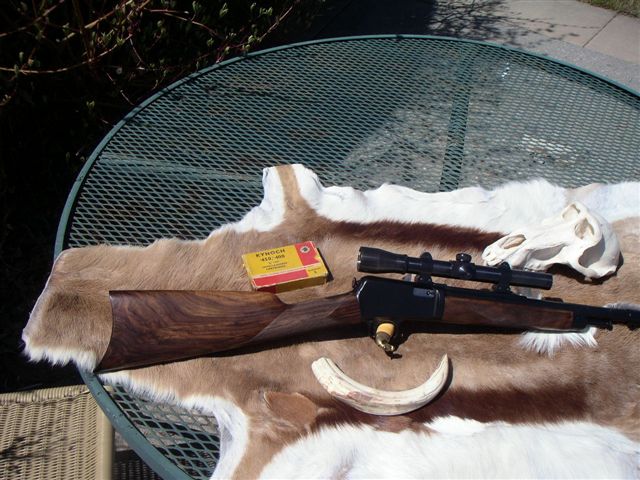 Rifle with scope, ammunition box, animal skull, and tusk on a fur rug on a metal table