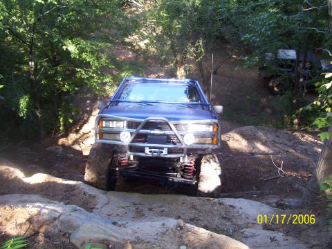 1978 Ford Bronco - A blue pickup truck is shown off-roading on a dirt and rock path with trees surrounding it.