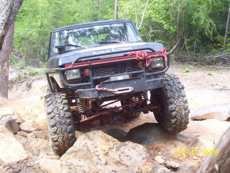 Black off-road vehicle with large tires navigating rocks, red grille guard, winch, and "ALASKAN EXTREME" text on windshield
