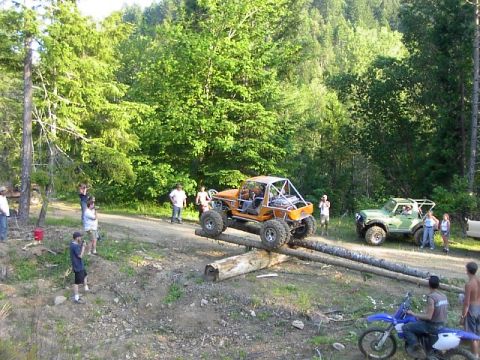1988 Ford Bronco - Two off-road vehicles navigate a log obstacle course with spectators observing the action.