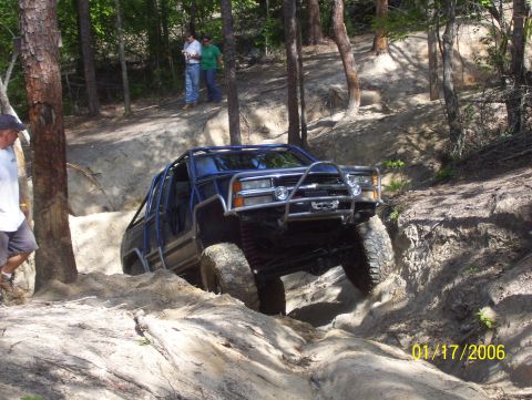 1978 Ford Bronco - A blue off-road vehicle with a prominent roll cage tackles a challenging dirt path in a wooded area.