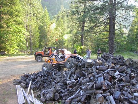 1988 Ford Bronco - Two off-road vehicles, an orange buggy and an orange SUV, are parked in a wooded area next to a large pile of cut logs.