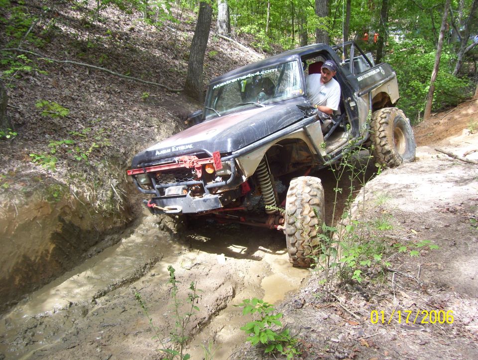 1978 Ford Bronco navigating a muddy trail, front end lowered into a rut, driver visible