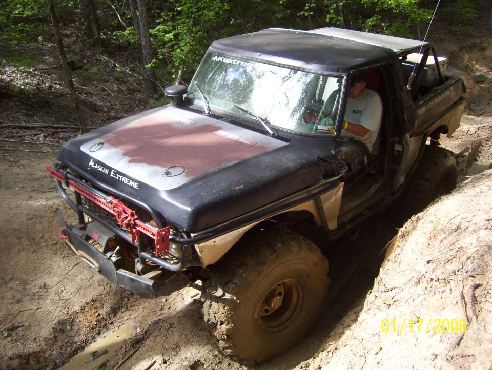 Modified pickup truck with off-road tires and roll cage navigating a dirt trail. Text on hood reads 'ALASKAN EXTREME'