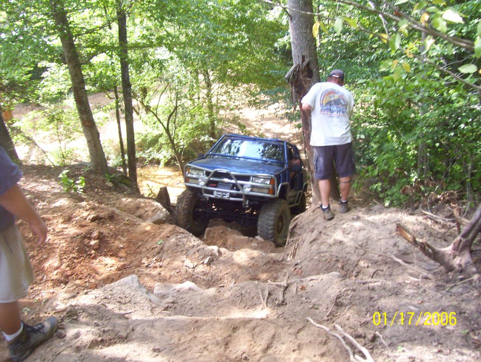 Blue off-road SUV with aftermarket bumper and large tires climbing a steep dirt hill with trees and a stream in the background