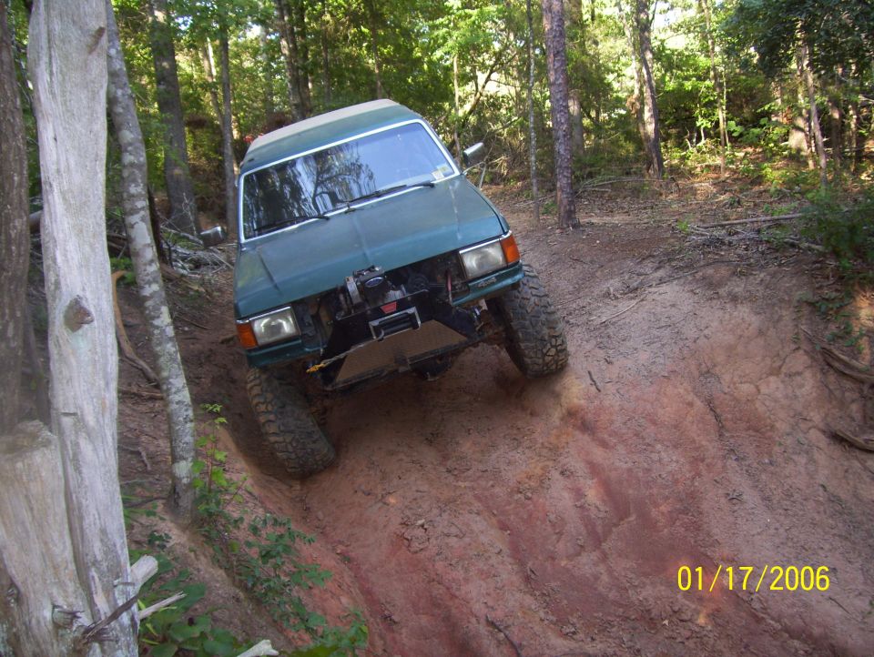 Green SUV with large tires going down a muddy hill in woods. Has a winch on front