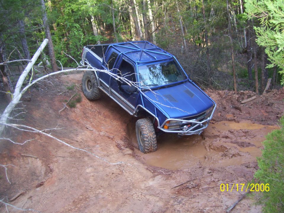 Blue custom off-road truck with roll cage driving through mud on a wooded trail