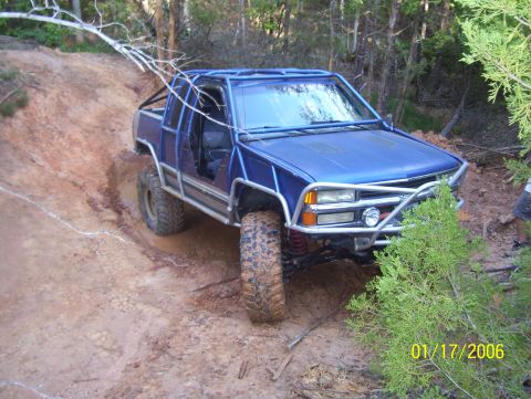 1978 Ford Bronco - A blue pickup truck with a roll cage navigates a muddy incline in a wooded area.