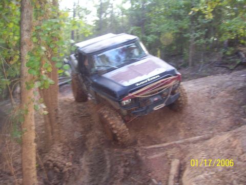 1978 Ford Bronco - A black off-road vehicle with a red hood is navigating a muddy trail in a wooded area. The vehicle has a winch and a roll cage visible on t…
