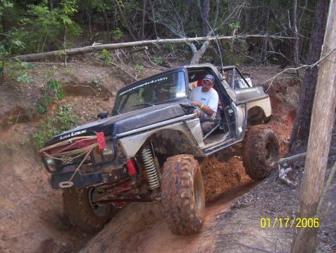 1978 Ford Bronco - A heavily modified black Ford Bronco is navigating a steep, muddy off-road trail with large, aggressive tires. The driver, wearing a baseba…