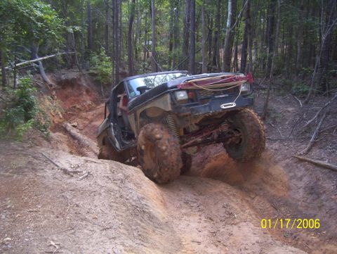1978 Ford Bronco - A lifted, mud-covered black Ford Bronco is navigating a steep, muddy off-road trail in a forest. The vehicle is angled upwards, with its fr…