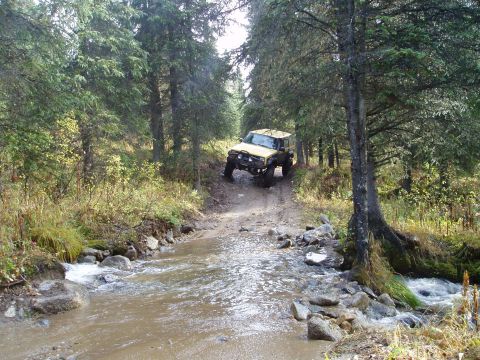 2000 Ford E350 - A yellow off-road vehicle tackles a challenging stream crossing on a dirt path surrounded by dense forest.