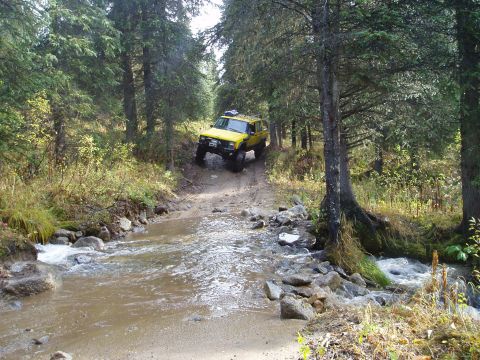 2000 Ford E350 - A yellow Jeep Cherokee navigates a stream crossing on a wooded trail, demonstrating off-road capability.