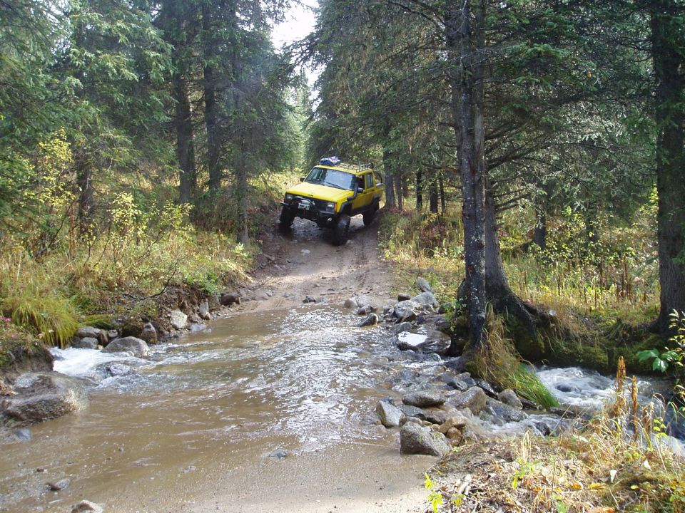 Yellow Jeep Cherokee driving through a stream on a dirt path in a forest
