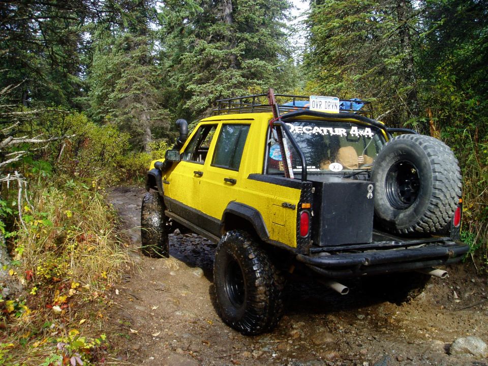 Yellow Jeep Cherokee XJ with off-road tires and spare tire on rear, driving on a dirt trail in a forest