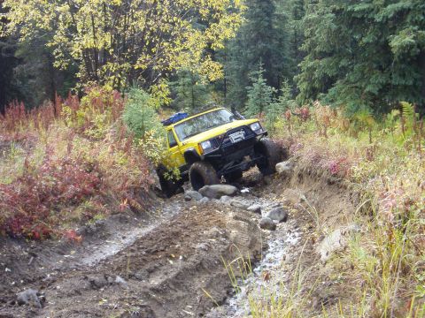 2000 Ford E350 - A yellow Jeep Cherokee tackles a challenging off-road trail with mud and rocks.