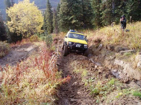 2000 Ford E350 - A yellow Jeep Cherokee navigates a challenging, muddy off-road trail in a wooded, mountainous area.