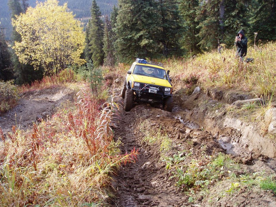 Yellow Jeep Cherokee climbing a muddy, rutted trail with a person filming in the background