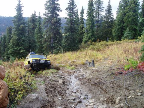 2000 Ford E350 - A yellow Jeep navigates a challenging, muddy off-road trail surrounded by dense forest.