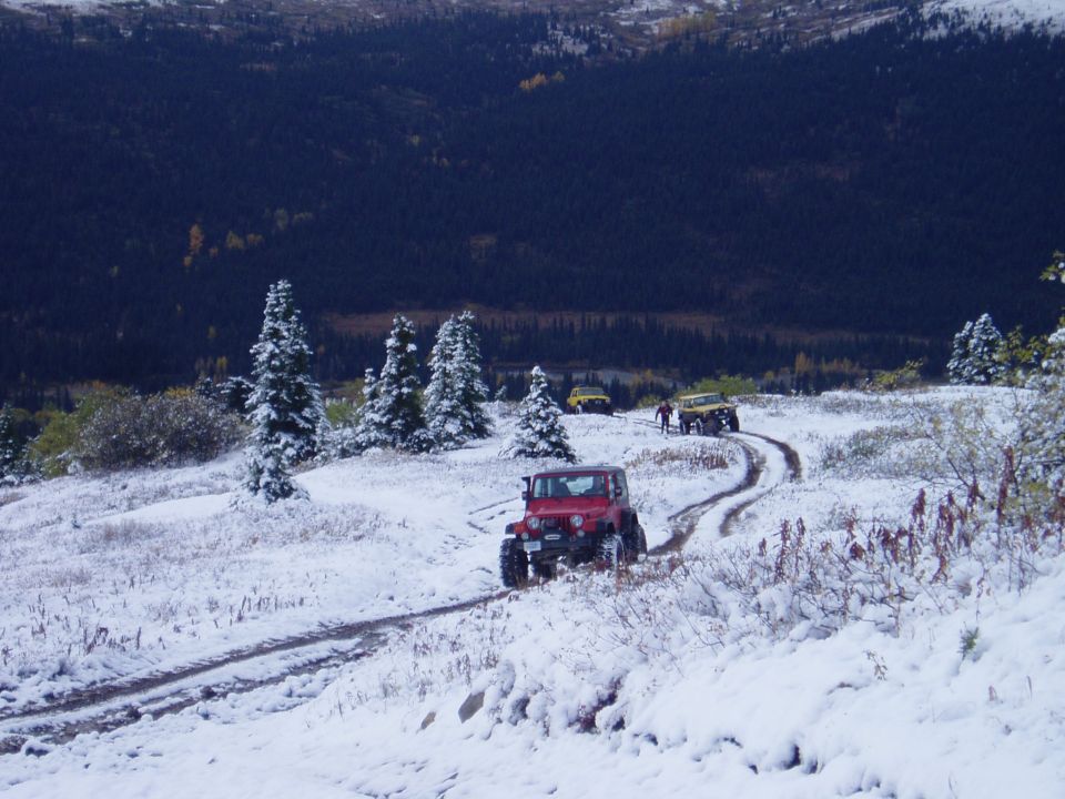Several Jeeps driving on a dirt road covered in snow, with a dark forest in the background