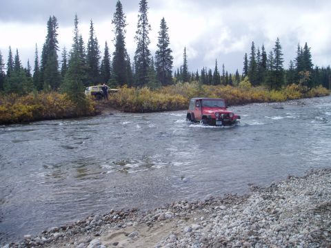 2000 Ford E350 - Two off-road vehicles navigate a rocky riverbed surrounded by trees under a cloudy sky.