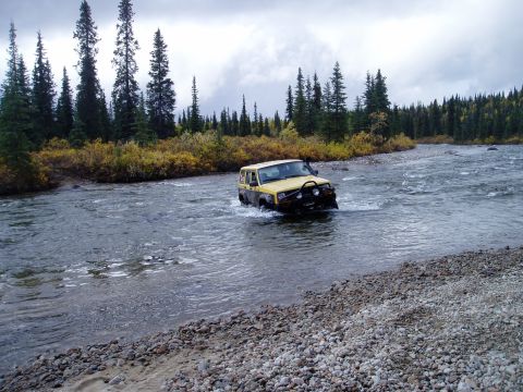 2000 Ford E350 - Yellow Jeep Cherokee XJ navigates a river crossing in a wooded area.