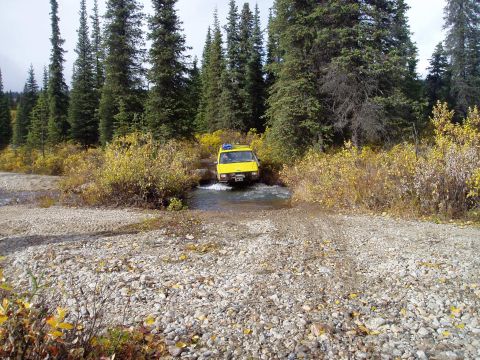 2000 Ford E350 - A yellow van navigates a water crossing on a gravel road surrounded by trees in autumn.