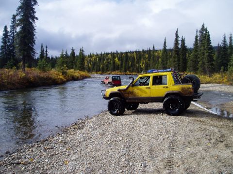 2000 Ford E350 - Two off-road vehicles navigate a shallow river surrounded by a forest of pine and autumn-colored trees.