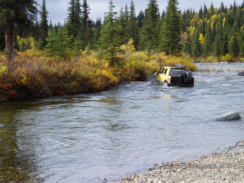 2000 Ford E350 - Yellow SUV navigates a river crossing surrounded by autumn foliage and evergreen trees.