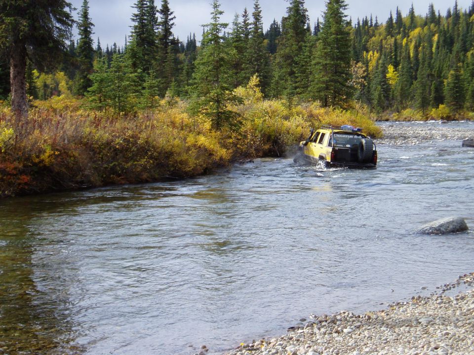 Yellow SUV with spare tire on back crossing a river, trees and brush on shore