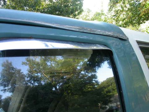 1990 Ford Bronco - The side window of a vehicle shows reflections of trees and sky. The trim appears weathered.