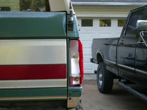1990 Ford Bronco - Rear view of a green and silver truck with a red stripe, parked next to a black pickup truck in front of a garage.