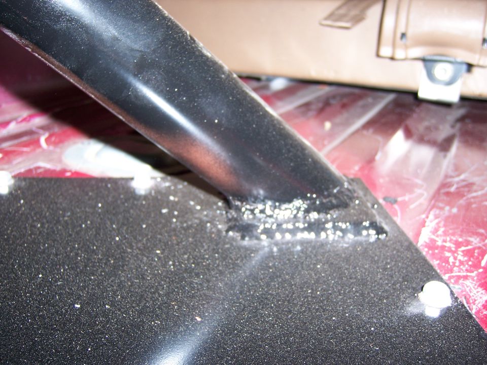 Black metal bracket with visible welds, bolted to a red surface, with a tan seat in the background