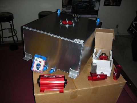 1998 Ford Crown Victoria - A bare aluminum fuel cell with blue AN bulkhead fittings and a red sender plate sits on a cardboard box, surrounded by red-anodized Aeromot…