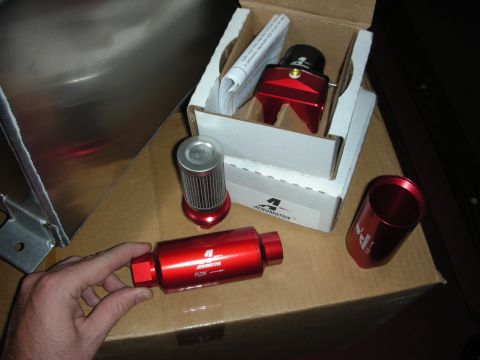 1998 Ford Crown Victoria - A hand steadies a red anodized Aeromotive inline fuel filter marked with a FLOW arrow on a cardboard box, with its disassembled housing and…