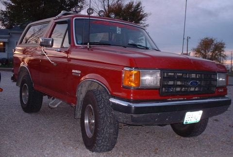 2006 Ford E350 - This red Ford Bronco is parked on a gravel surface with trees in the background.