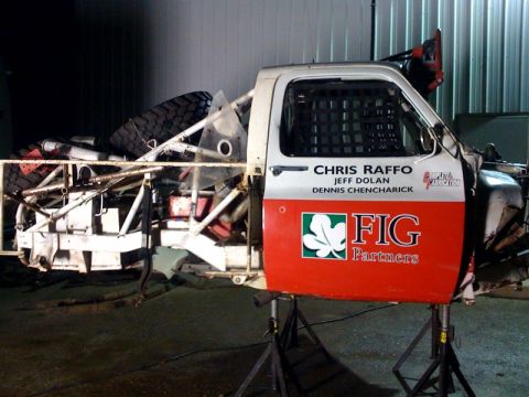 1990 Ford Bronco - red and white off-road race truck cab sits on jack stands in a shop, lettered with FIG Partners and driver names Chris Raffo, Jeff Dolan an…