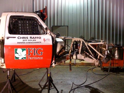 1990 Ford Bronco - red and white race truck cab and its exposed tube-frame chassis rest on jack stands inside a metal-walled shop.