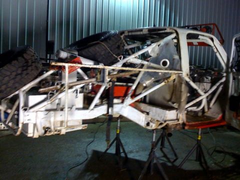1990 Ford Bronco - stripped white off-road race truck tube chassis sits on multiple jack stands against a corrugated metal wall.