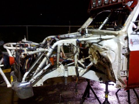1990 Ford Bronco - partially built white off-road race truck chassis with exposed suspension and wiring sits on jack stands outdoors at night.