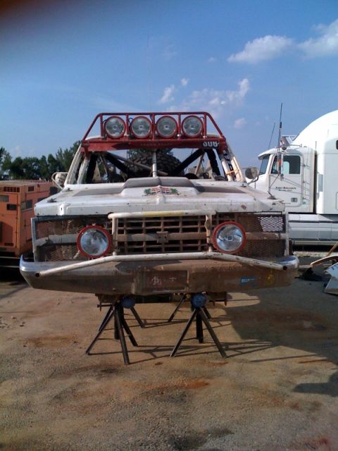 1990 Ford Bronco - weathered off-road race truck with a roof-mounted light bar and round headlights sits on jack stands in a dirt yard beside a semi truck.