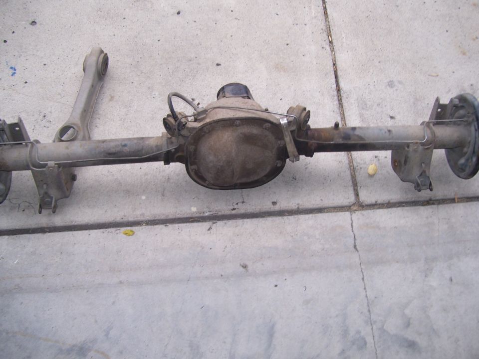 Removed Ford solid rear axle assembly with differential cover and brake lines lying on concrete