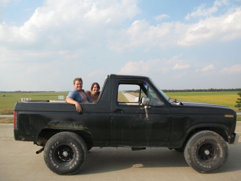 1985 Ford Bronco - Two people ride in a black Ford Bronco on a rural road with fields on either side.