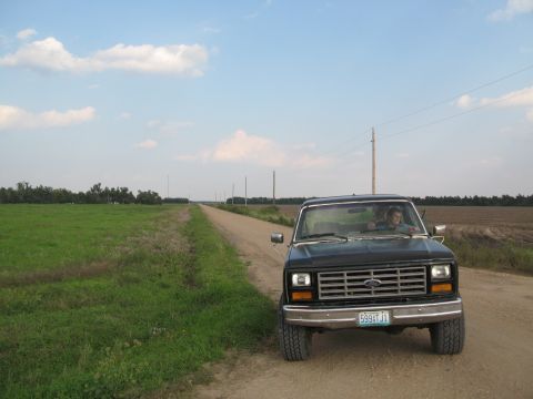 1985 Ford Bronco - A black Ford F-Series truck is parked on a dirt road with a field to the left and farmland to the right under a cloudy sky.
