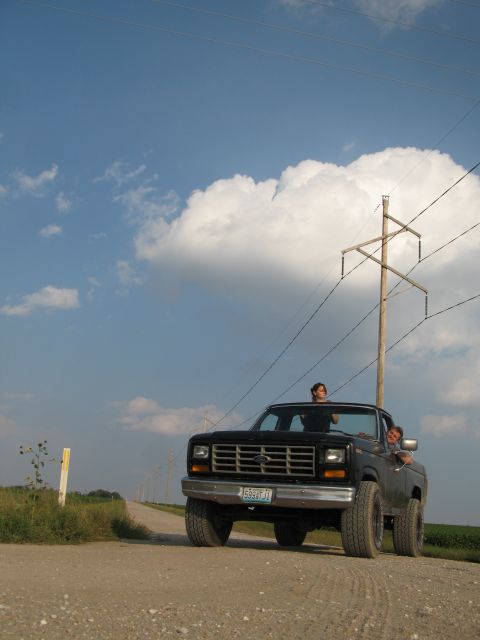 1985 Ford Bronco - A black pickup truck with two people inside drives down a dirt road under a cloudy sky.