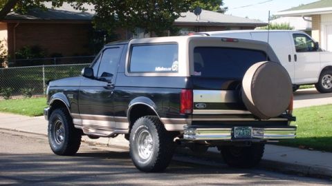 1996 Ford Bronco - The rear view of a two-tone Ford Bronco shows its spare tire and a white van parked nearby.
